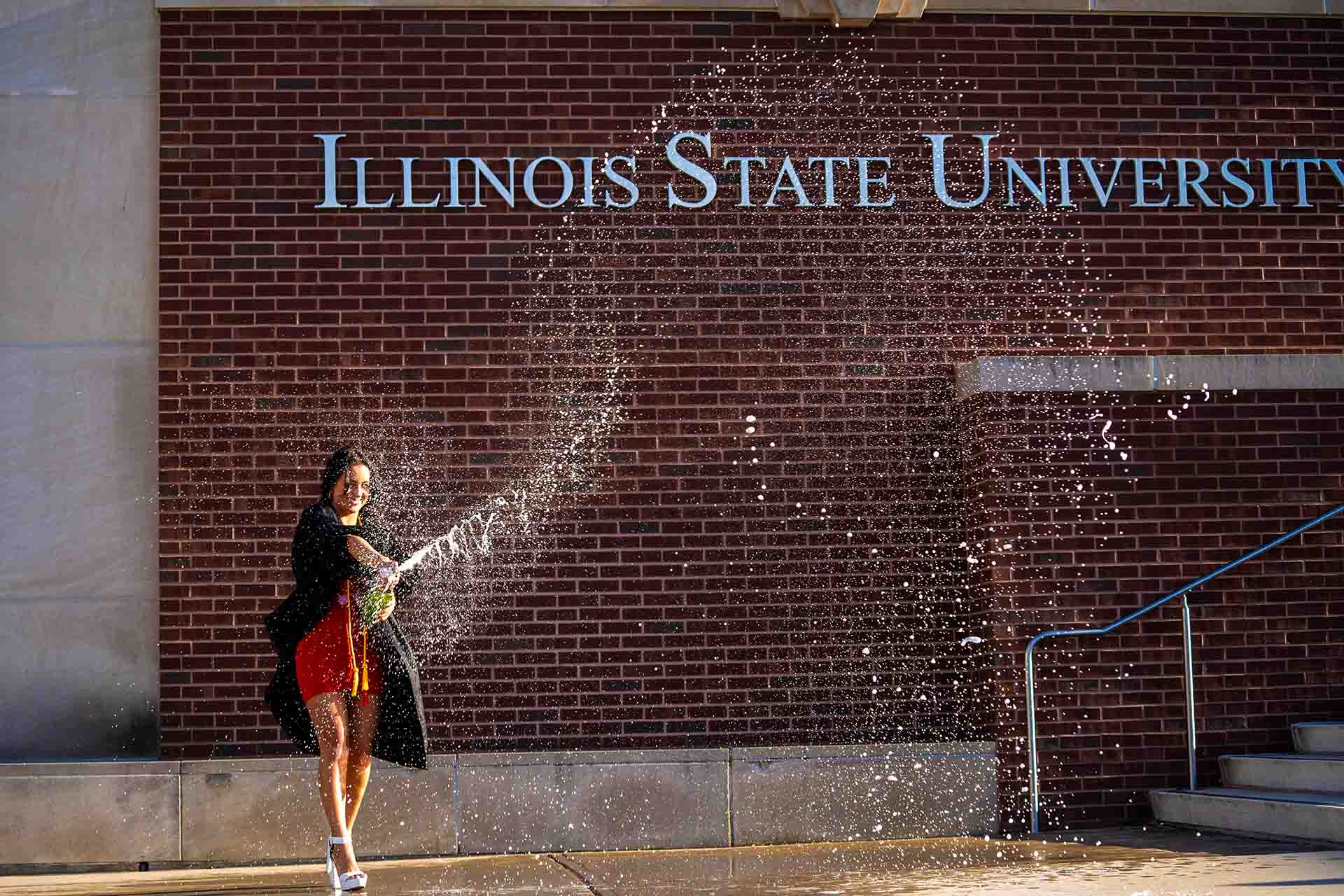 Graduate popping champagne in front of the Illinois State University wall